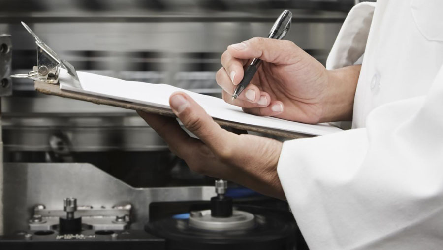  A factory worker wearing a lab coat is writing on a clipboard while inspecting machinery.